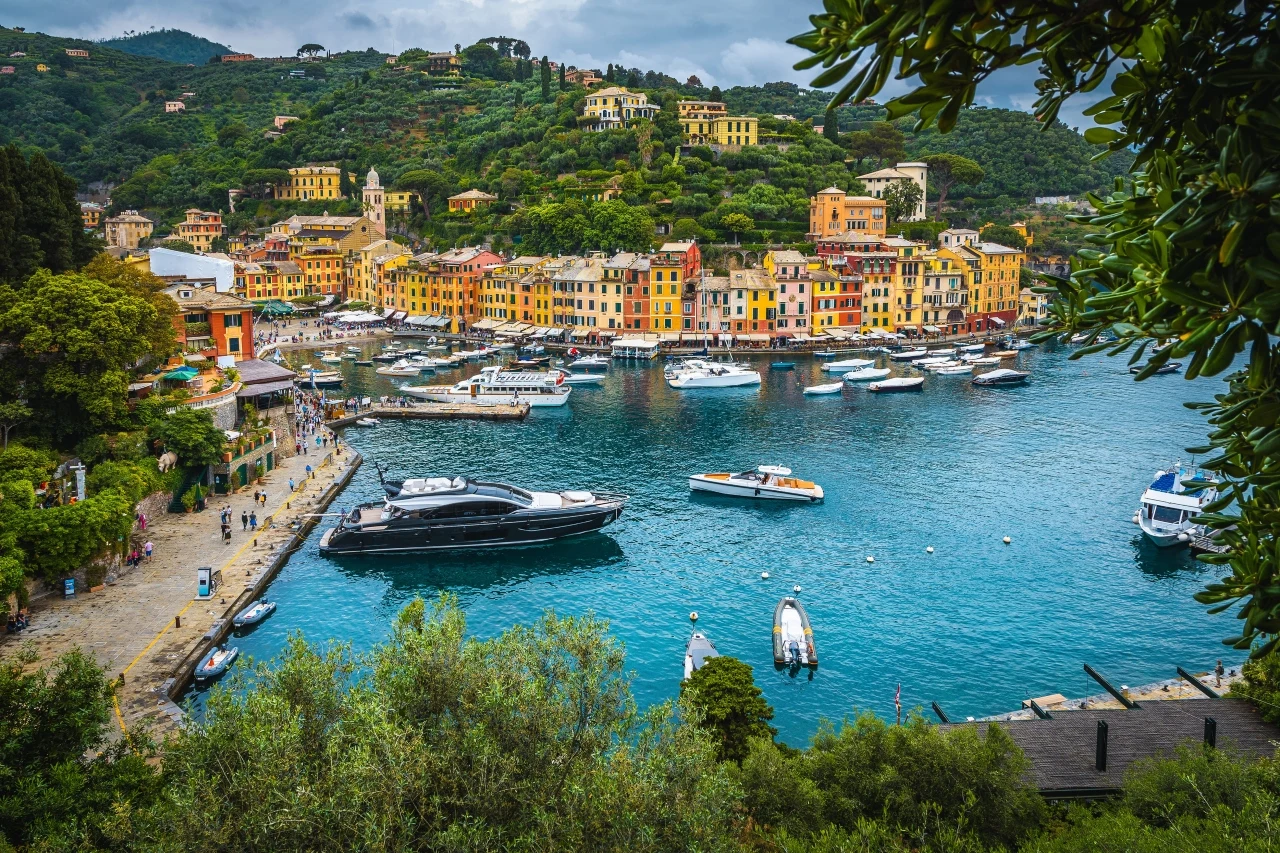 Portofino Kolorowe budynki wznoszące się nad mariną pełną jachtów i łodzi, otoczone zielonymi wzgórzami porośniętymi drzewami i krzewami.