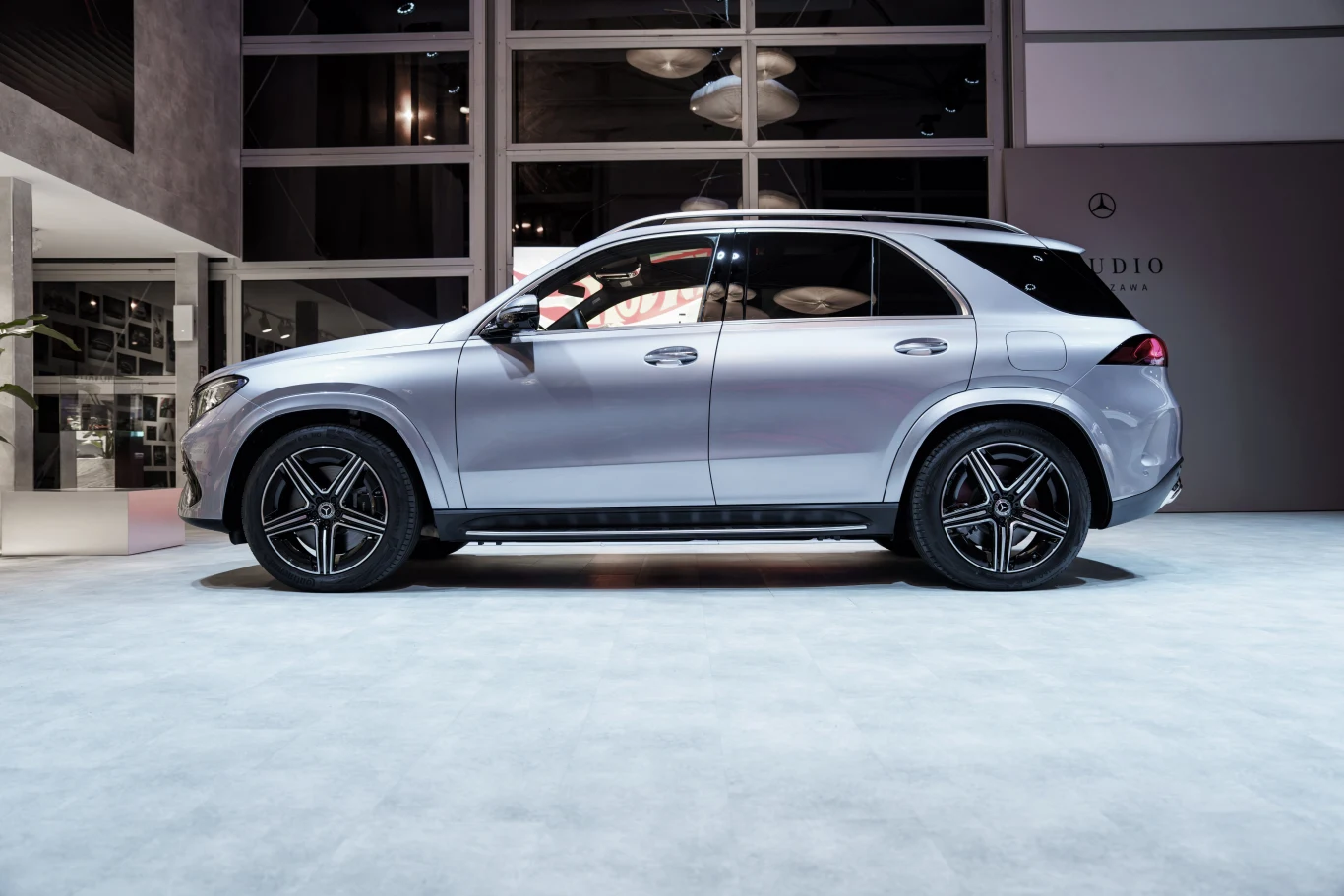 Srebrny SUV marki Mercedes-Benz z przyciemnianymi szybami i charakterystycznymi pięcioramiennymi felgami, ustawiony w jasnym, nowoczesnym wnętrzu wystawowym.