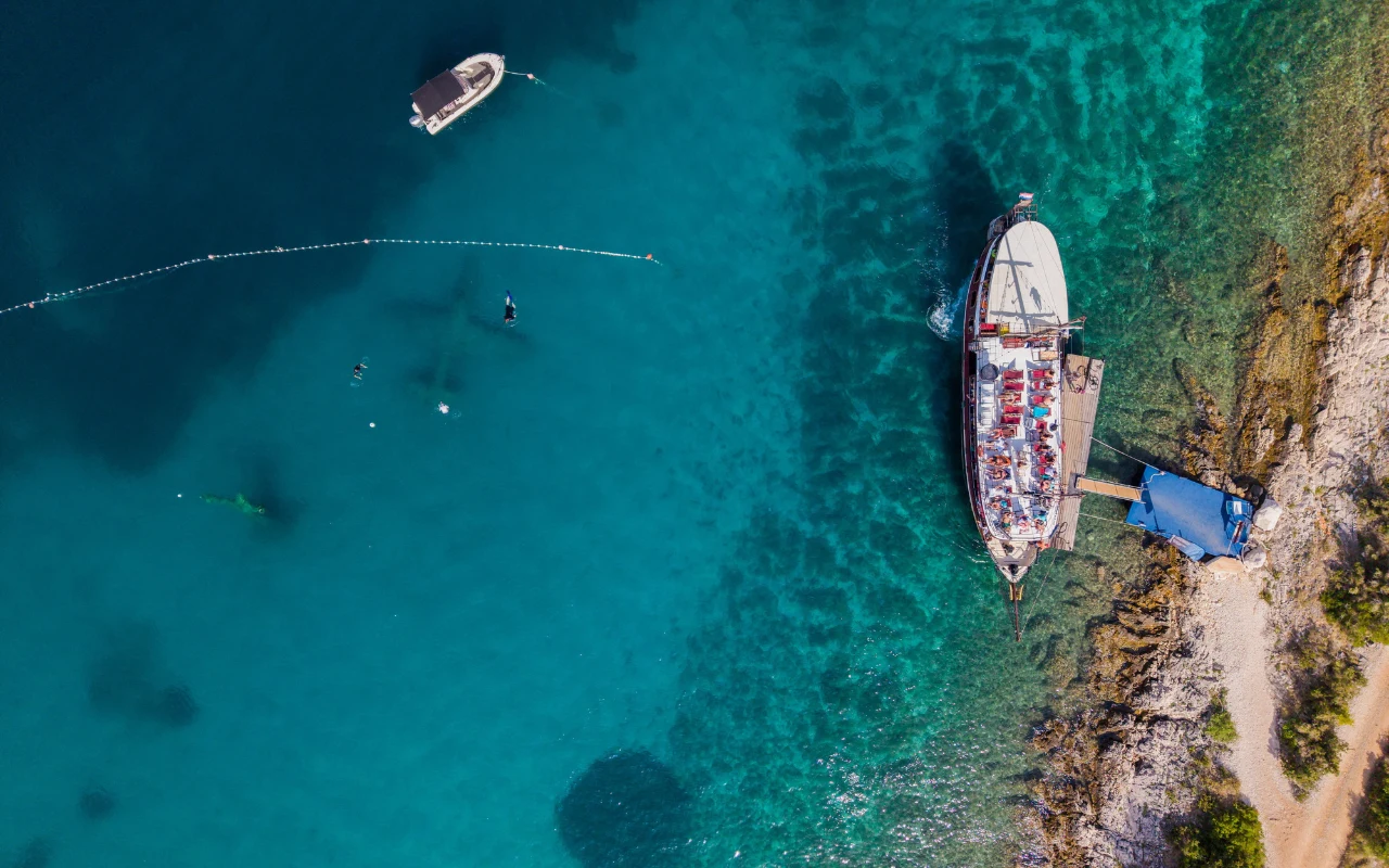 W Underwater Musem Croatia eksponaty znajdują się pod wodą Duża łódź zacumowana przy kamienistym brzegu, kilka osób pływających w przejrzystej, niebieskiej wodzie oraz nieduża łódź dryfująca nieopodal.