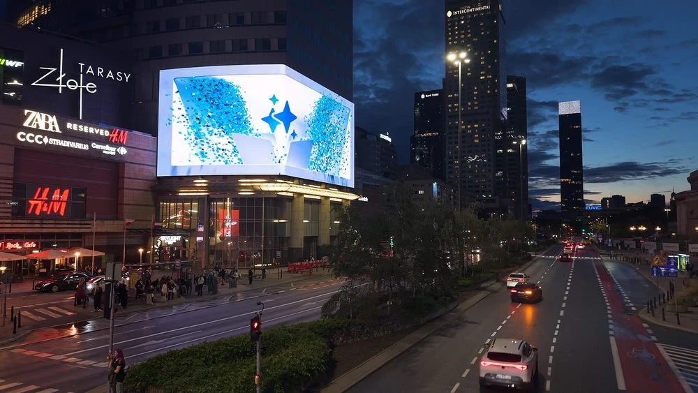 Zajawka Galaxy Unpacked wyświetlana na WARSAW Stage. Duży ekran LED na fasadzie galerii Złote Tarasy wyświetla animację ze świecącą gwiazdą, poniżej widoczny ruch uliczny, piesi i samochody, a w tle nowoczesne wieżowce Warszawy oświetlone światłami miasta po zmierzchu.