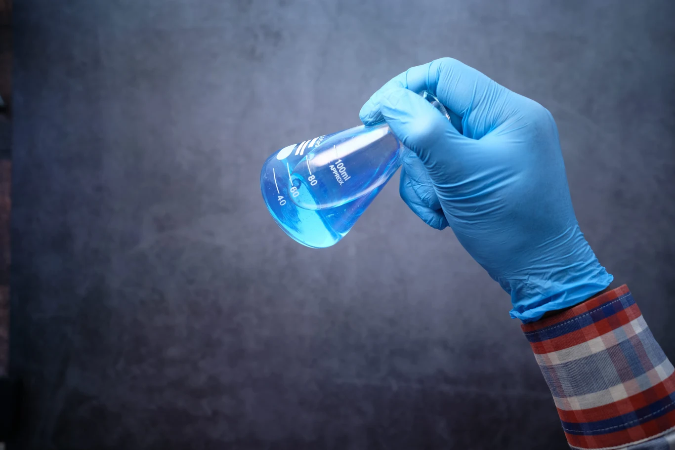 Niemieccy naukowcy opracowują nowe techniki usuwania "wiecznych chemikaliów". Ręka w niebieskiej rękawiczce ochronnej trzyma szklaną kolbę z niebieskim płynem laboratoryjnym.