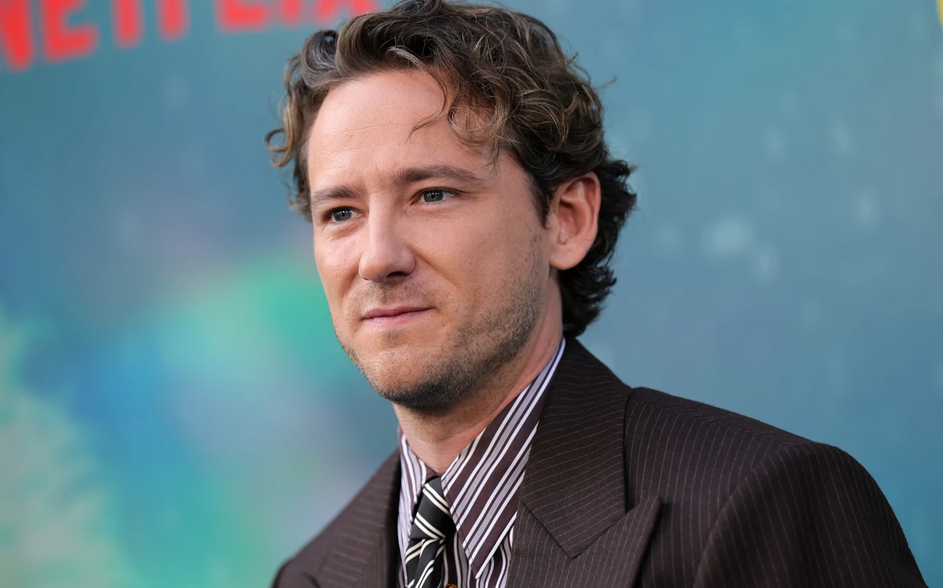Lewis Pullman Lewis Pullman w brązowym garniturze i koszuli w paski na tle rozmytego plakatu z logo Netflix.