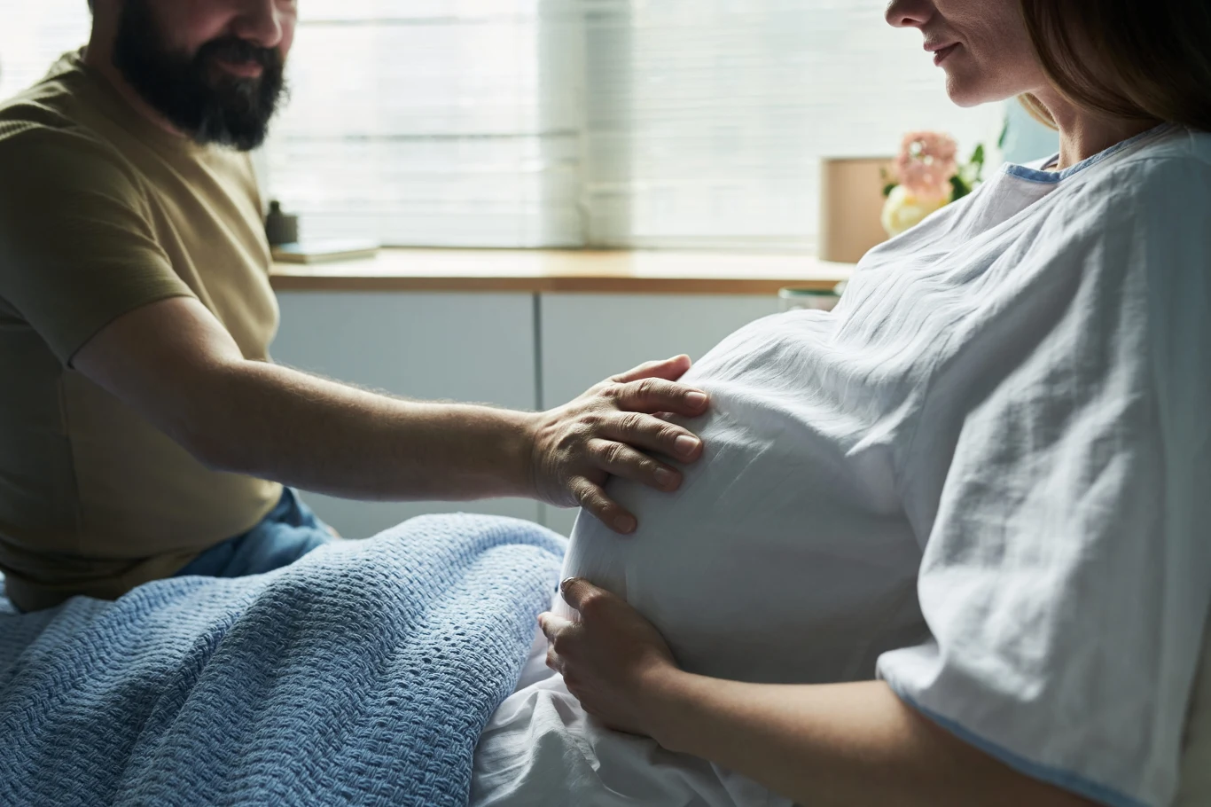 Mężczyzna delikatnie dotyka brzucha kobiety w zaawansowanej ciąży, oboje siedzą blisko siebie w jasnym pomieszczeniu.