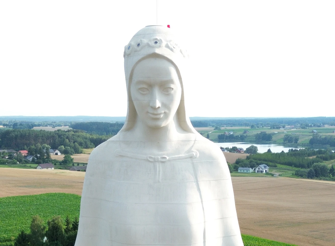 Zbliżenie na górną część pomnika Matki Boskiej w Konotopiu Biała, monumentalna figura kobiety z koroną na głowie góruje nad rozległymi polami i wsią, w tle widoczne są lasy oraz jezioro.
