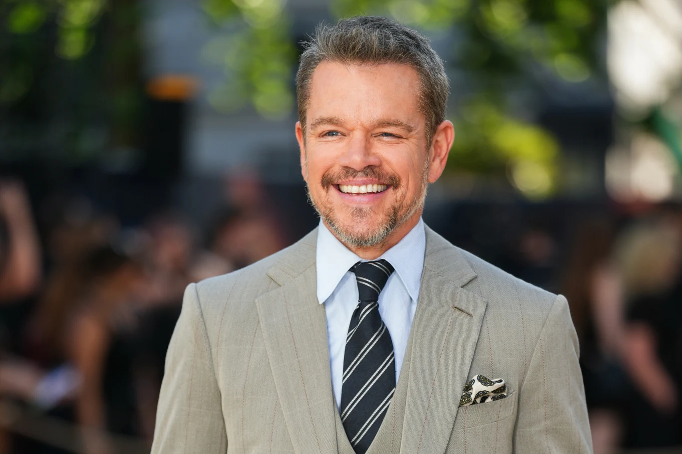 Matt Damon Matt Damon w jasnoszarym garniturze, białej koszuli i krawacie w paski na zielonym tle.