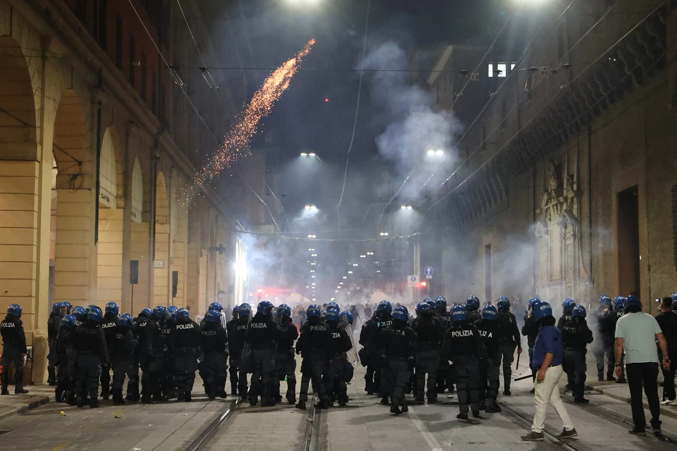 Oddział policji ubrany w pełne opancerzenie stoi w zwartej formacji na ulicy nocą, nad głowami funkcjonariuszy wybuchają race, a atmosfera jest napięta i zadymiona.