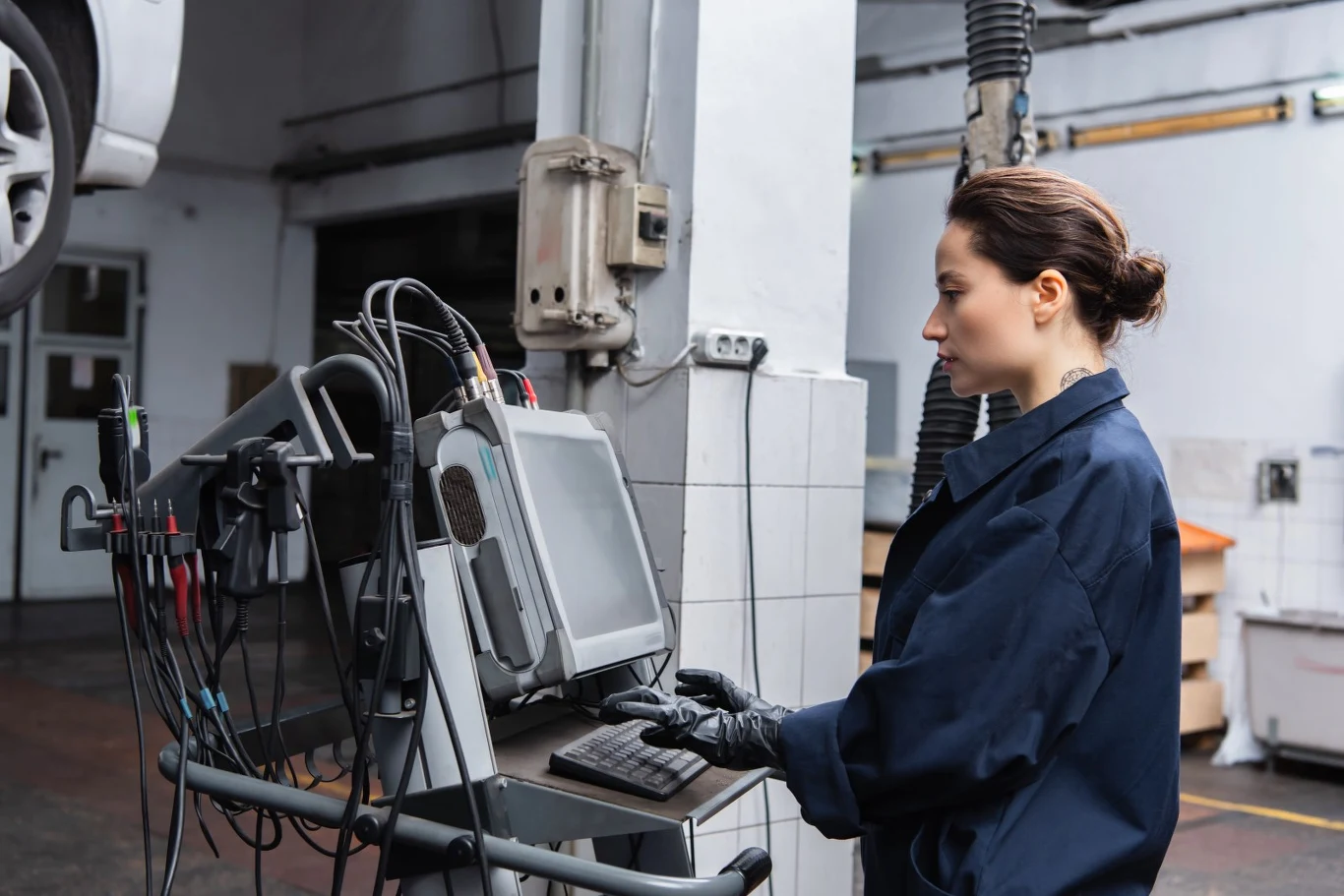 Kobieta w kombinezonie roboczym obsługuje komputer diagnostyczny w warsztacie samochodowym, wokół niej widoczne są przewody i urządzenia techniczne.