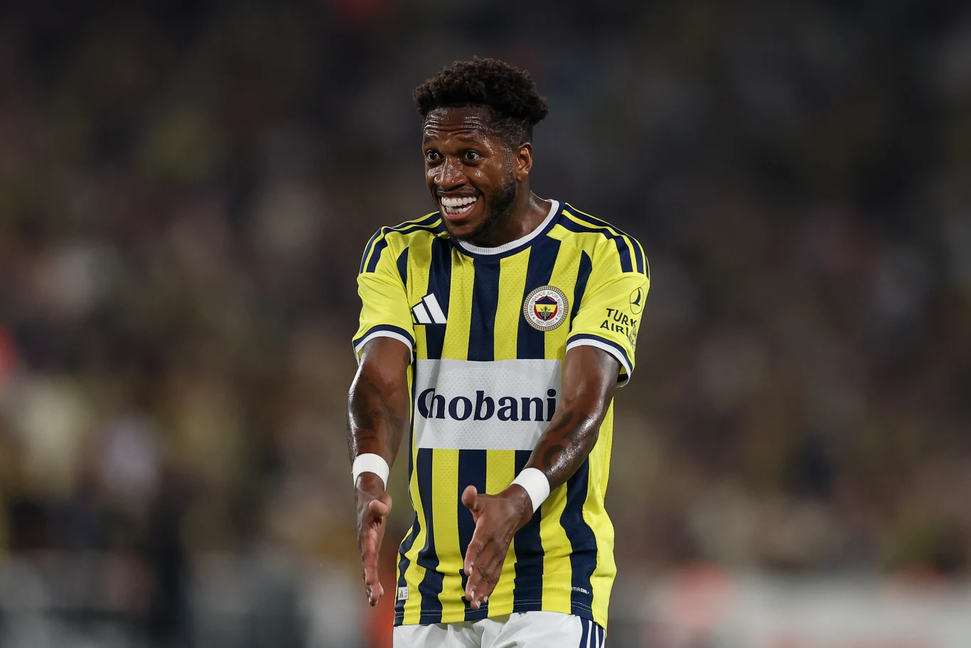 Piłkarz w żółto-niebieskiej koszulce Fenerbahçe, wykonujący gesty rękami, z wyrazem silnych emocji na twarzy, podczas meczu na stadionie.