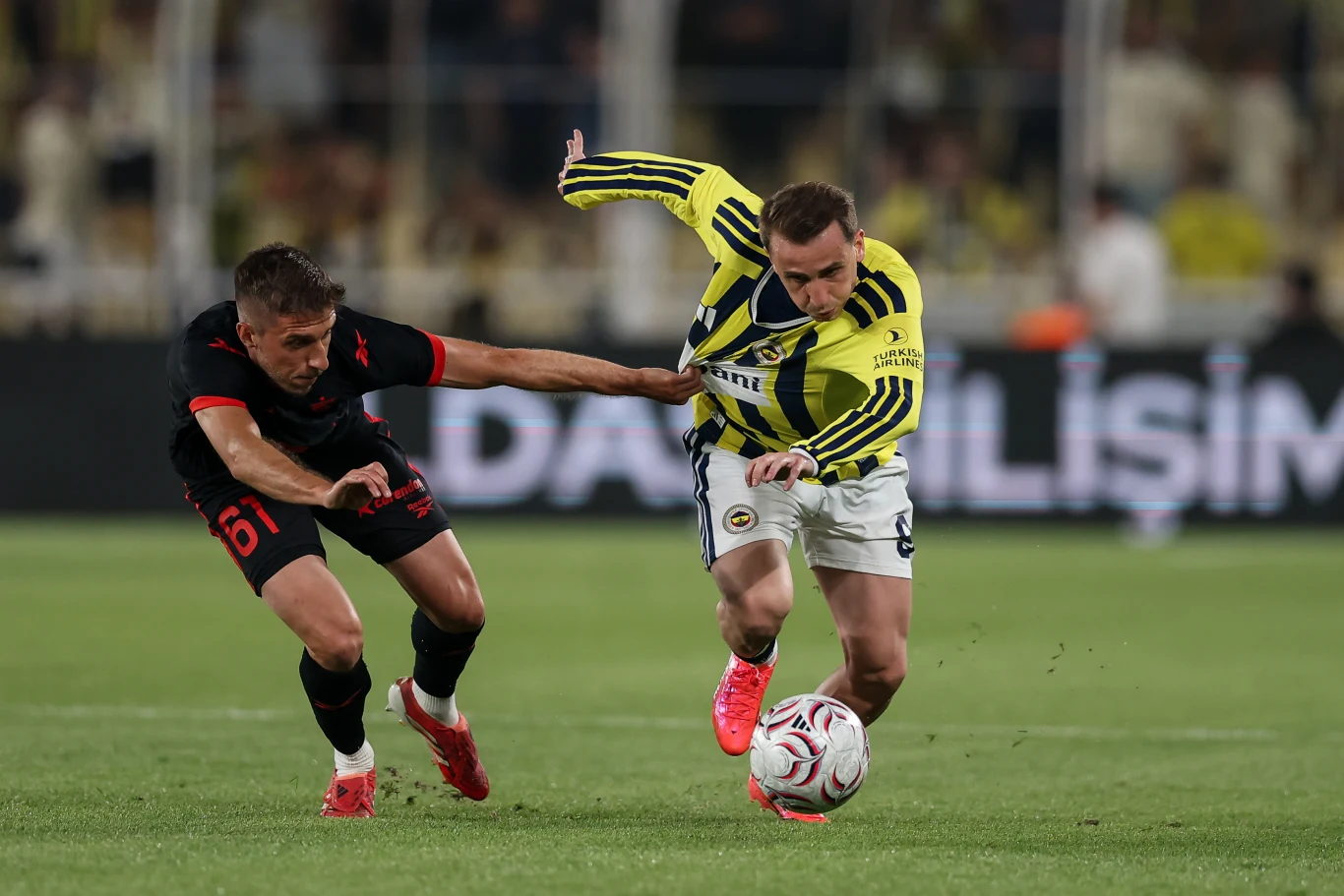 Intensywna walka dwóch piłkarzy o piłkę podczas meczu, jeden z zawodników w żółto-granatowej koszulce Fenerbahçe próbuje przejąć kontrolę nad piłką, drugi gracz w czarno-czerwonym stroju sięga ręką, by go powstrzymać.