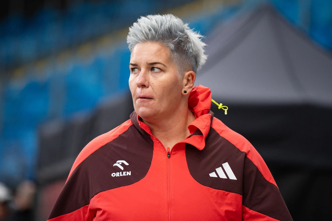 Osoba z krótkimi, siwymi włosami w czerwono-czarnej sportowej kurtce z logo Orlen i adidas, patrząca w bok, na tle rozmytych niebieskich trybun stadionu.