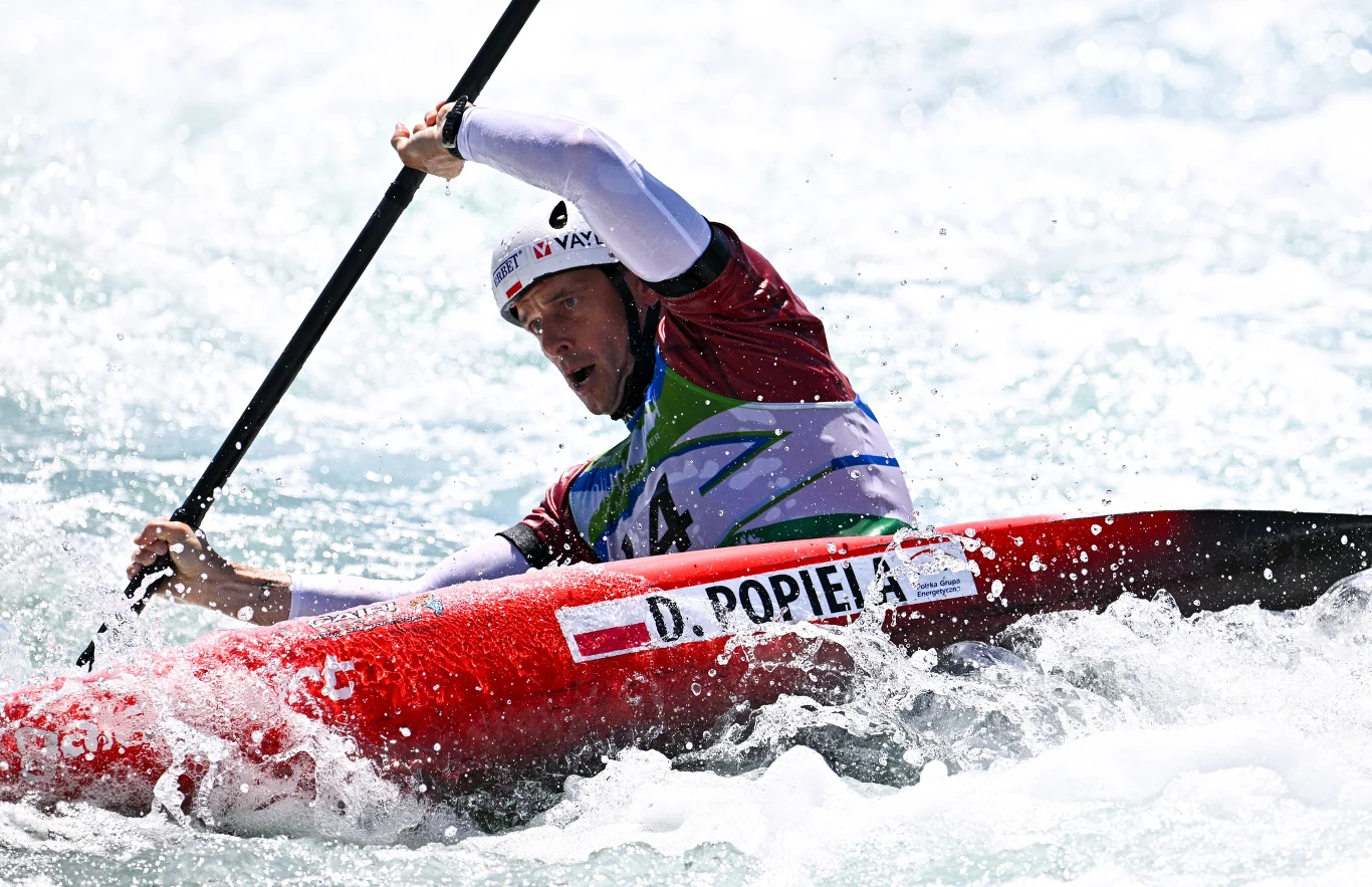 Kajakarz w sportowym stroju dynamicznie płynie kajakiem po wzburzonej wodzie, energicznie manewrując wiosłem.