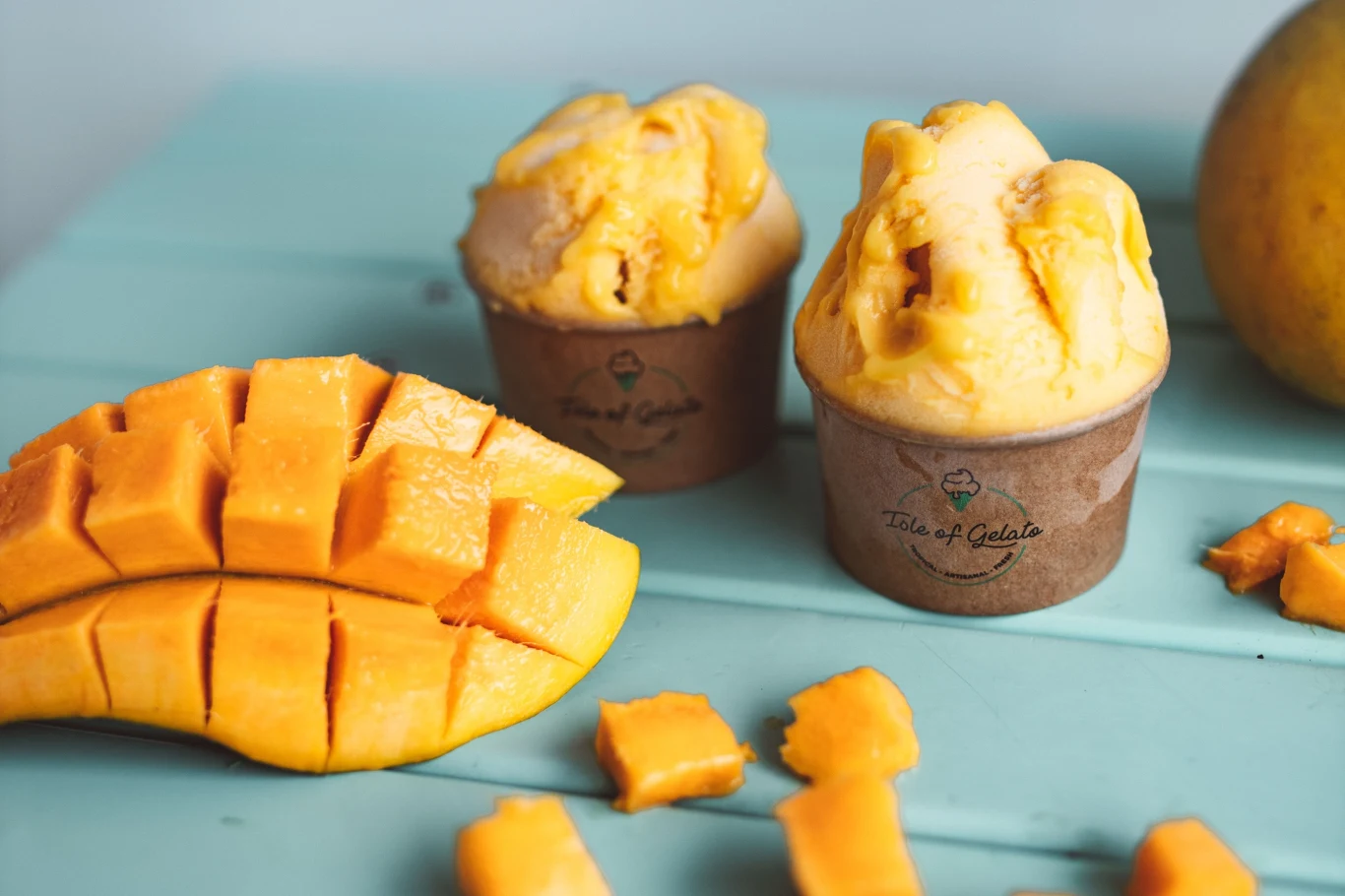 Lody z mrożonego mango i serka wiejskiego to hit na redukcji Dwie porcje lodów z mrożonego mango i serka wiejskiego w papierowych kubeczkach obok kostek mango na turkusowym blacie.