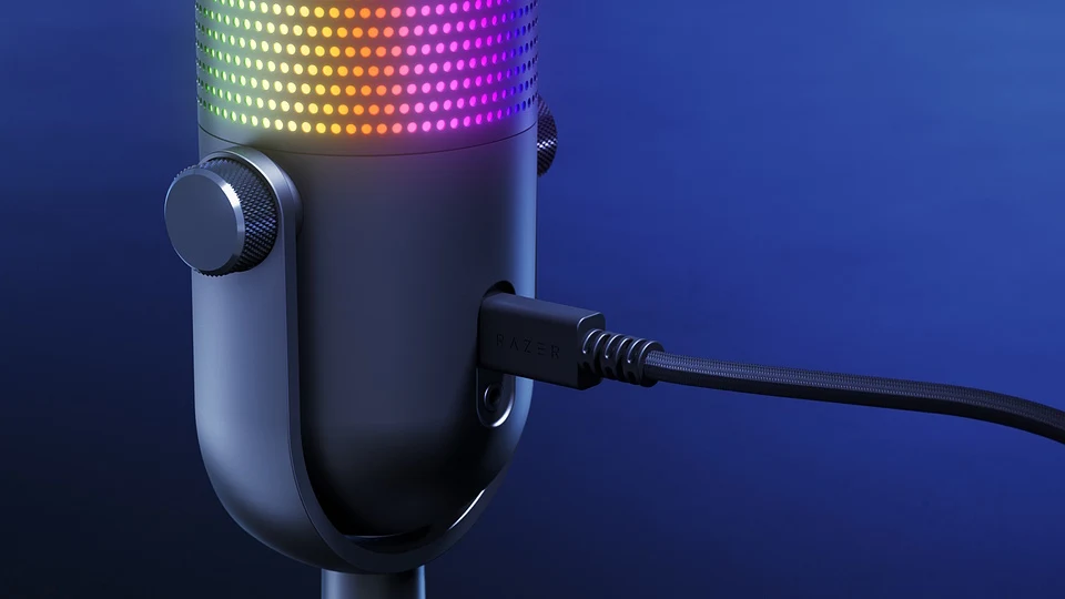 Czarny mikrofon z kolorowym podświetleniem LED, połączony przewodem USB na tle niebieskiego gradientu.