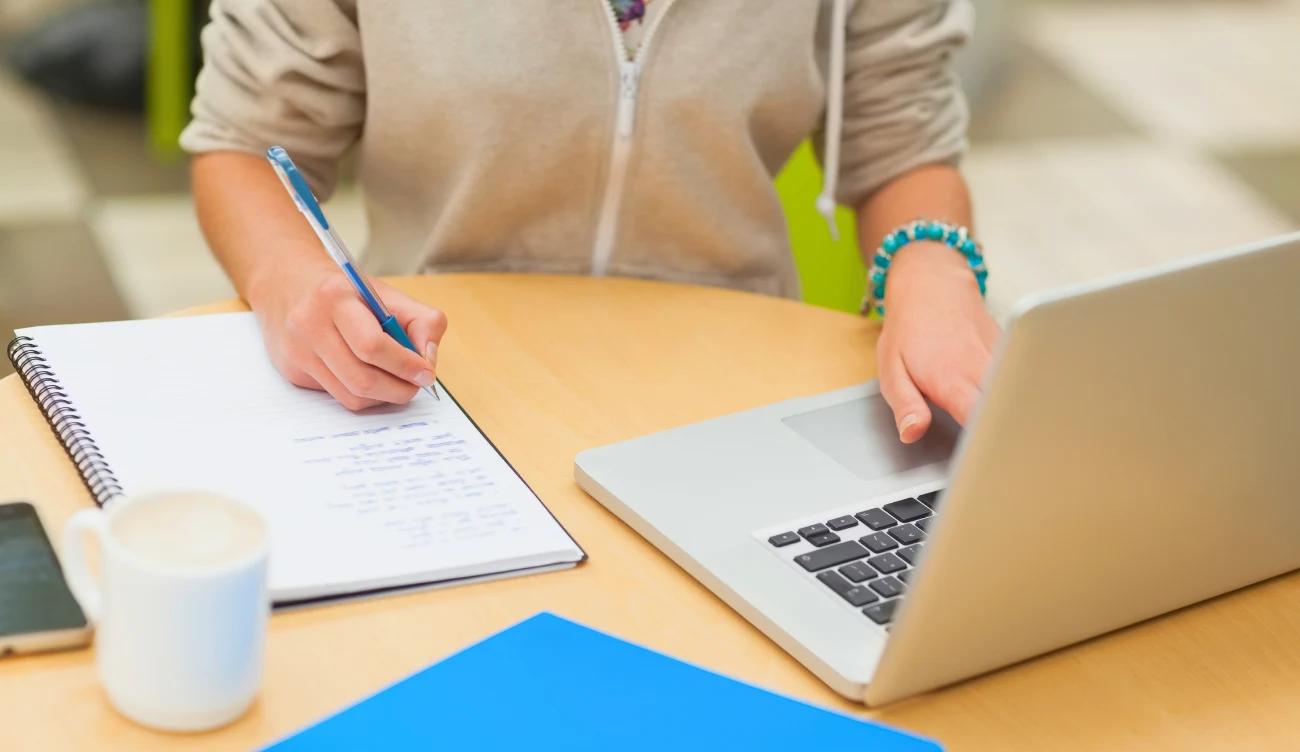 Stypendia dla studentów 2026 Osoba w jasnej bluzie przy okrągłym stole pisze w notesie i korzysta z laptopa, na blacie teczka i smartfon.