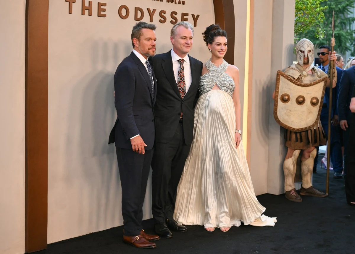 Trzech elegancko ubranych dorosłych pozuje razem na tle dekoracji wydarzenia filmowego, obok nich stoi postać w stroju antycznego wojownika z tarczą i hełmem.