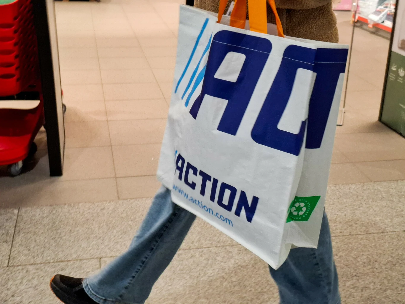 Osoba niosąca dużą torbę zakupową z logo sklepu Action, widoczne jeansy oraz pomarańczowe i białe uchwyty torby.