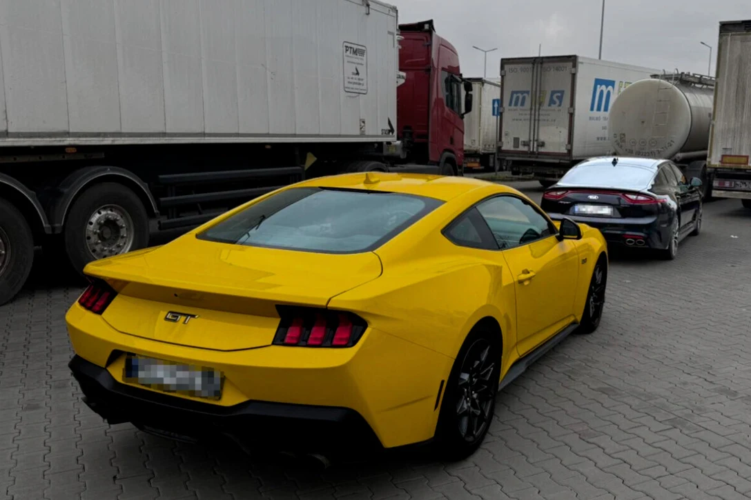 Jaskrawożółty Ford Mustang GT zaparkowany na placu obok ciemnego sedana i dużych ciężarówek.