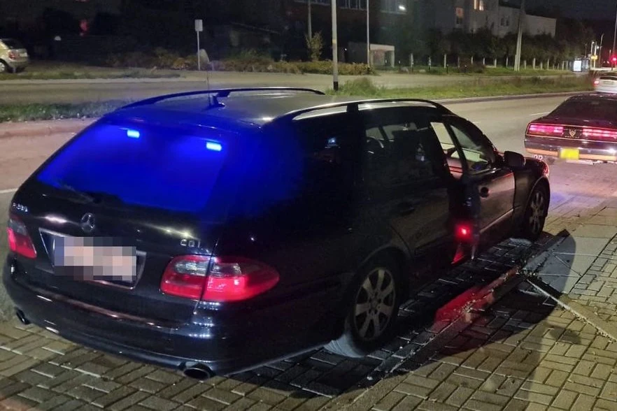 Czarny samochód osobowy marki Mercedes z otwartymi drzwiami na poboczu drogi, ze świecącymi niebieskimi światłami w środku oraz stojący za nim inny pojazd.