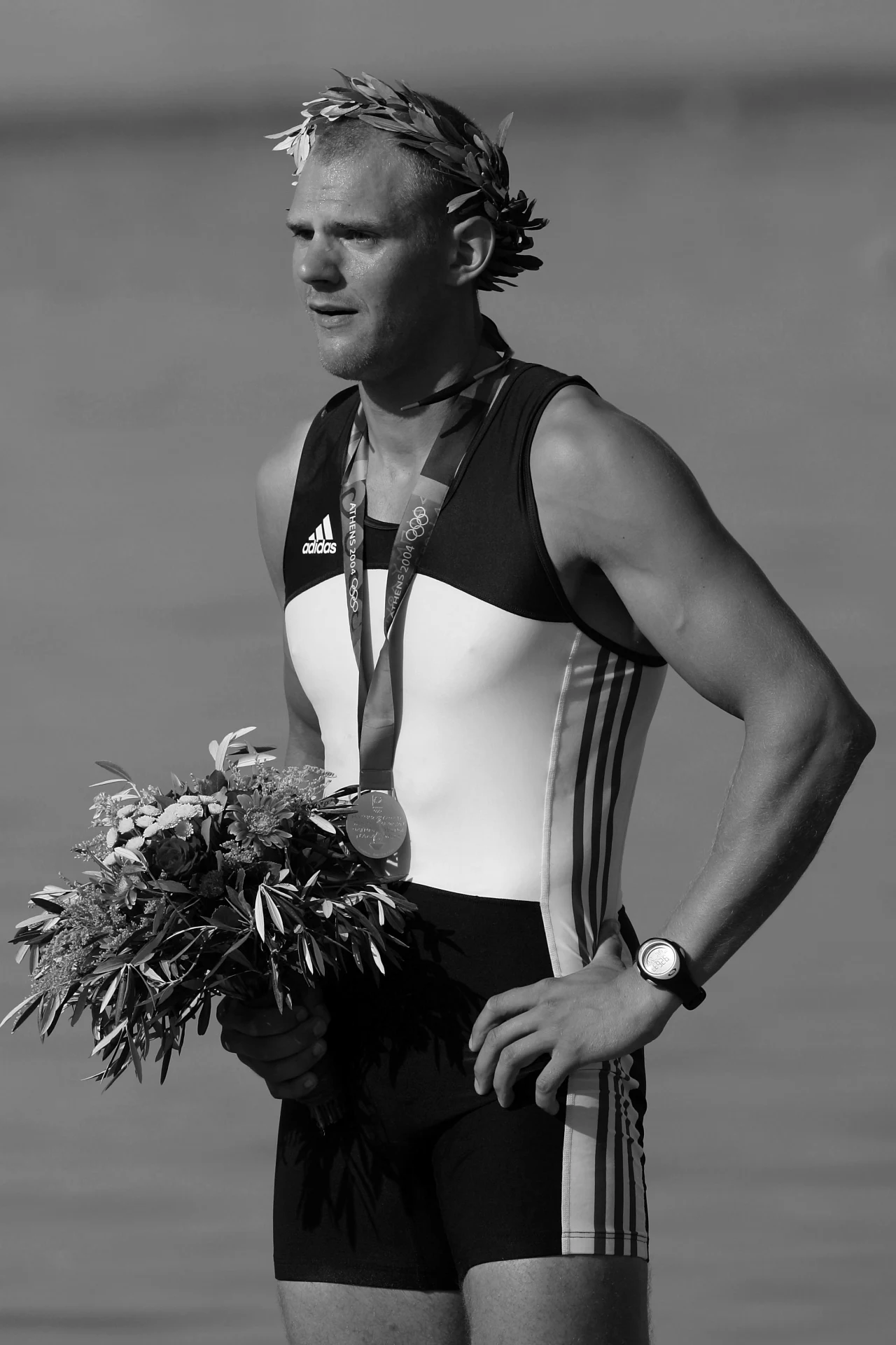 Olaf Tufte Sportowiec w stroju startowym z medalem na szyi, wieńcem laurowym na głowie i bukietem kwiatów w dłoni, stojący na tle rozmytej tafli wody.