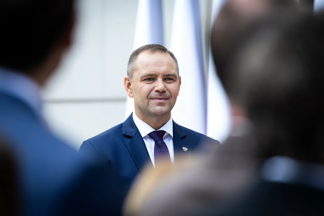 Mężczyzna w garniturze z krawatem stoi w centrum kadru, uśmiechając się lekko, w tle widoczne białe flagi oraz niewyraźne sylwetki innych osób.