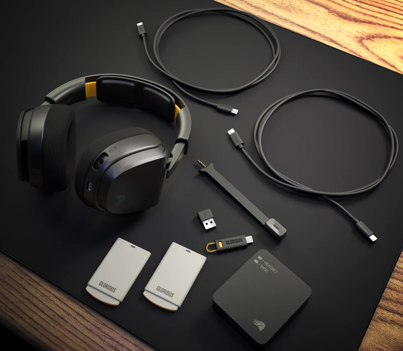 Glorious GHS Wireless InfinitePlay Słuchawki nauszne wraz z akcesoriami: dwie ładowarki, dwa kable USB-C, dwa adaptery USB oraz czarne etui, wszystko ułożone na czarnej powierzchni obok drewnianego blatu.