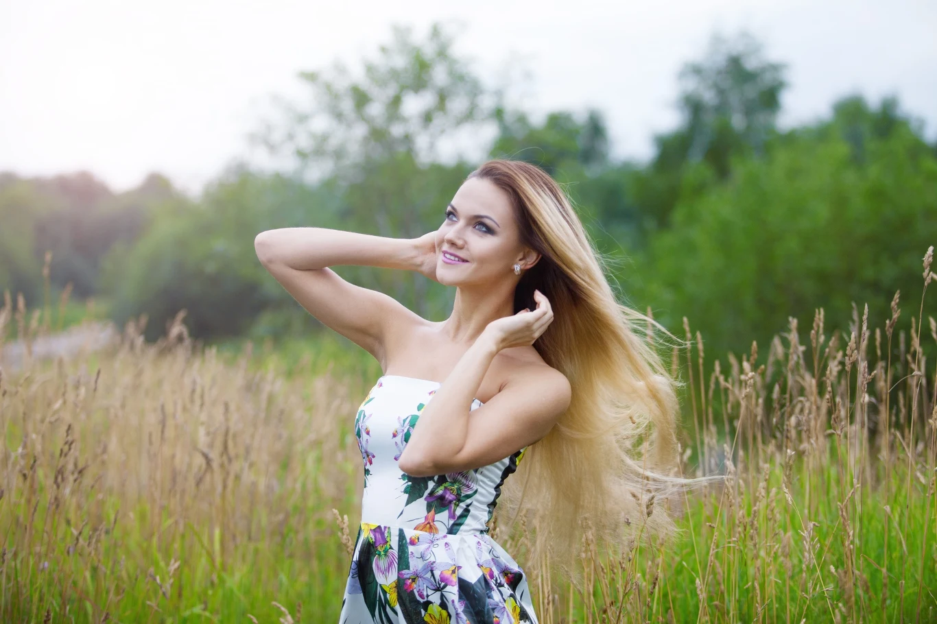 Uśmiechnięta kobieta w sukience w kwiaty stoi pośród wysokich traw, unosząc ręce do długich blond włosów, na tle zieleni i rozmytych drzew.