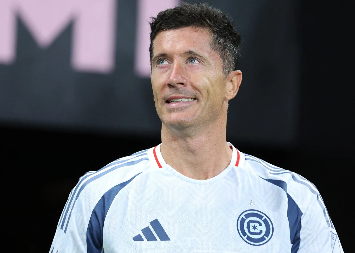 Mężczyzna w sportowej koszulce z logo drużyny, stojący na tle ciemnego rozmytego otoczenia, z lekko uśmiechniętą twarzą unoszącą się ku górze.