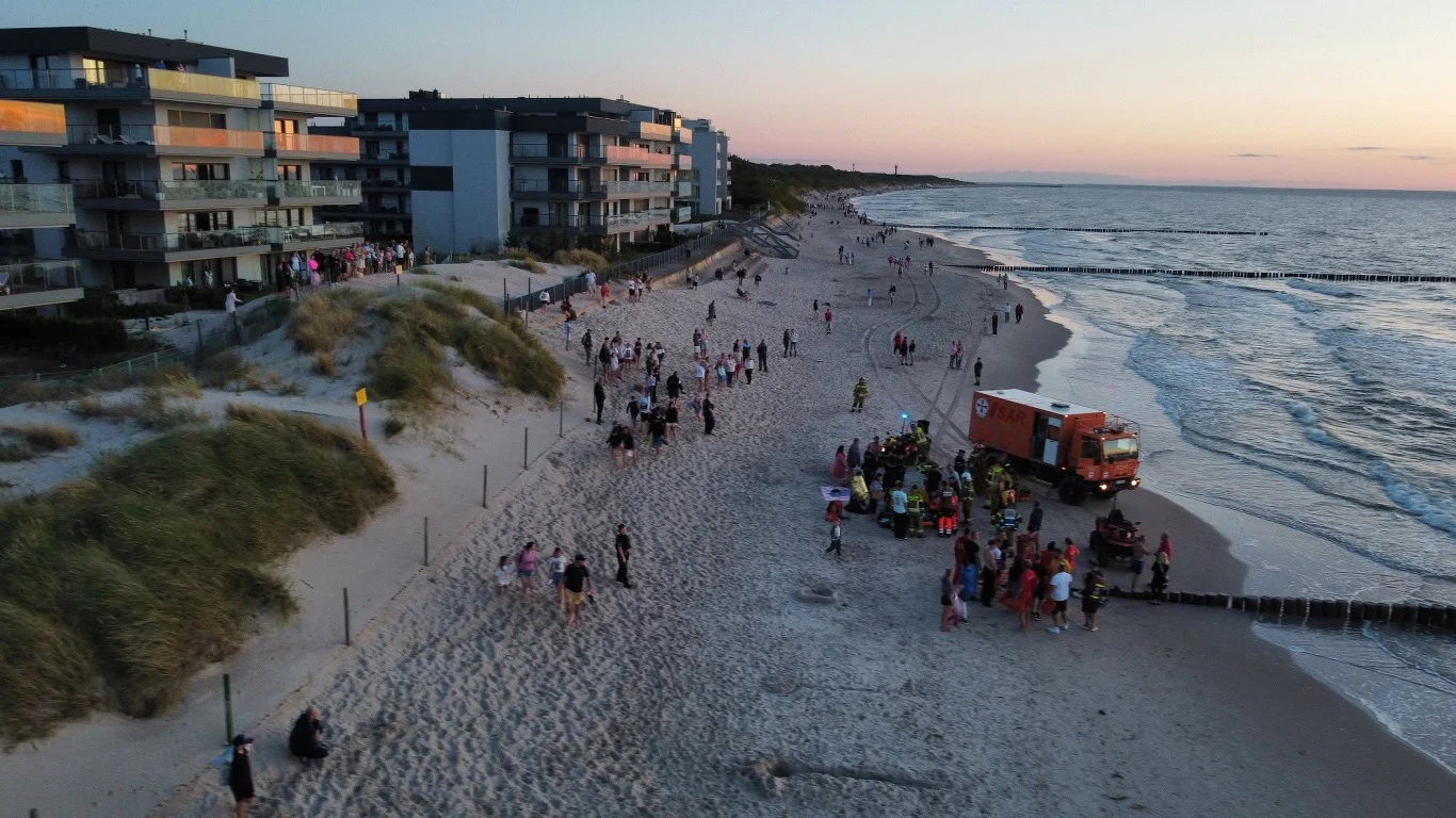 Tłum ludzi zgromadzony wokół czerwonego pojazdu ratowniczego na szerokiej, piaszczystej plaży przy linii nowoczesnych apartamentowców, z widokiem na spokojne morze o zachodzie słońca.