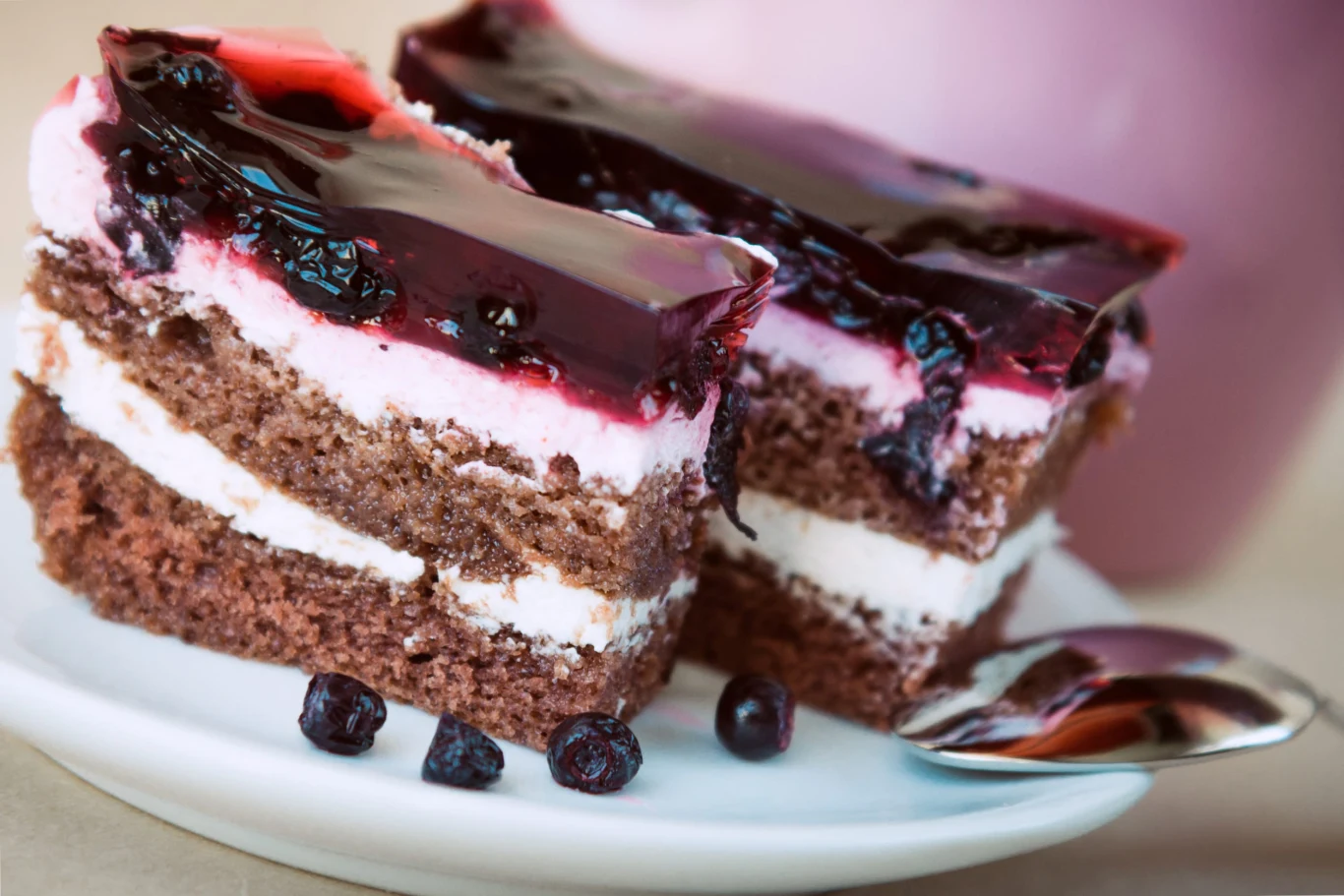 Slice of blueberry pie with yoghurt Dwa kawałki czekoladowo-jagodowego placka z kremem i galaretką na białym talerzu, obok łyżeczka i jagody.