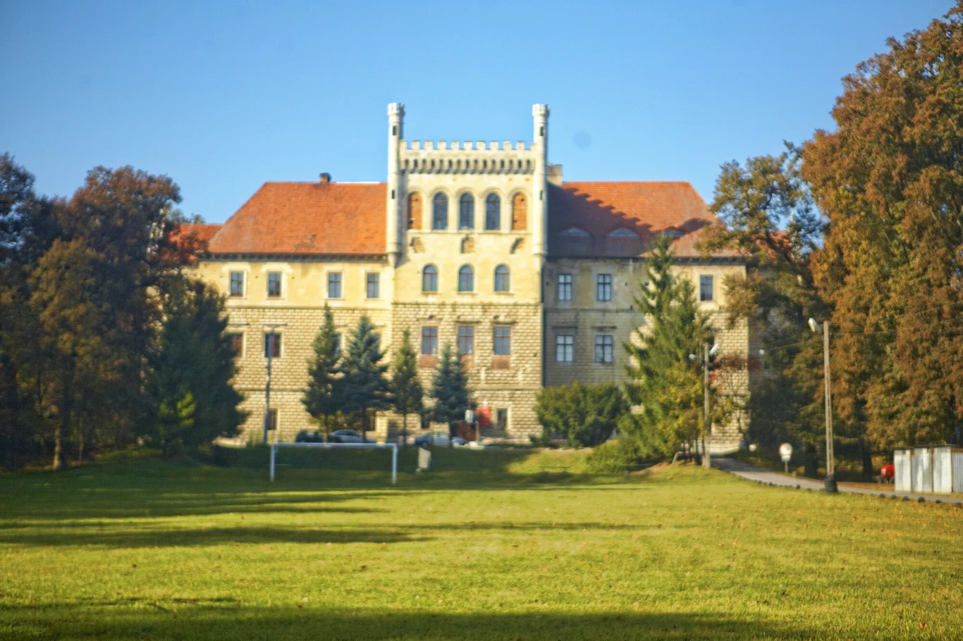 Duży zabytkowy pałac z jasnej cegły z czerwonym dachem, otoczony zielonymi drzewami i rozległym trawnikiem na pierwszym planie.