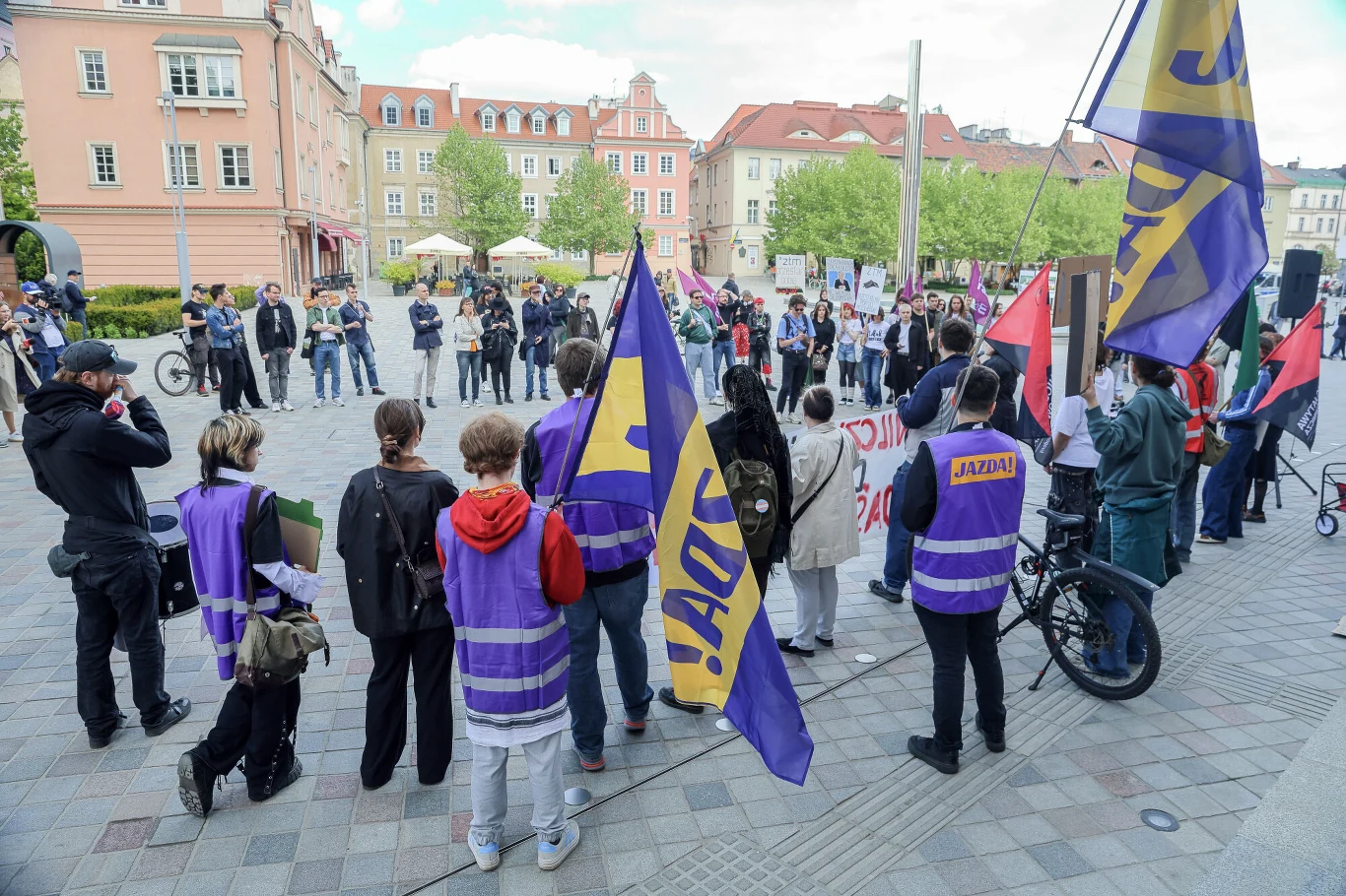 Grupa ludzi z fioletowymi kamizelkami i flagami z napisem „Jazda” zgromadzona na miejskim placu podczas manifestacji lub protestu, w tle kamienice i kilka osób obserwujących wydarzenie.