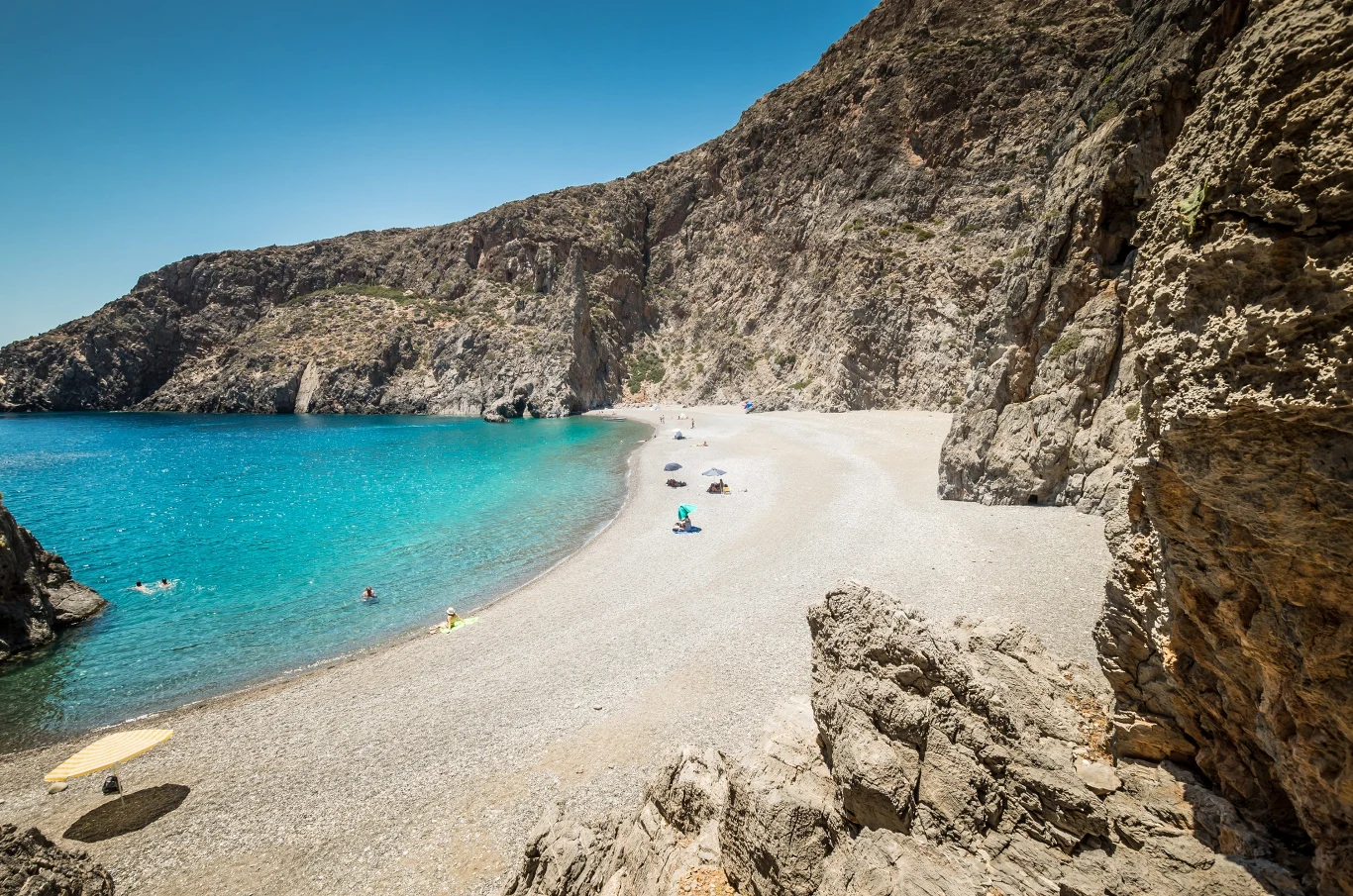 Malownicza zatoka z turkusową wodą, szeroką żwirową plażą i kilkoma plażowiczami odpoczywającymi pod parasolami, otoczona wysokimi, skalistymi klifami.