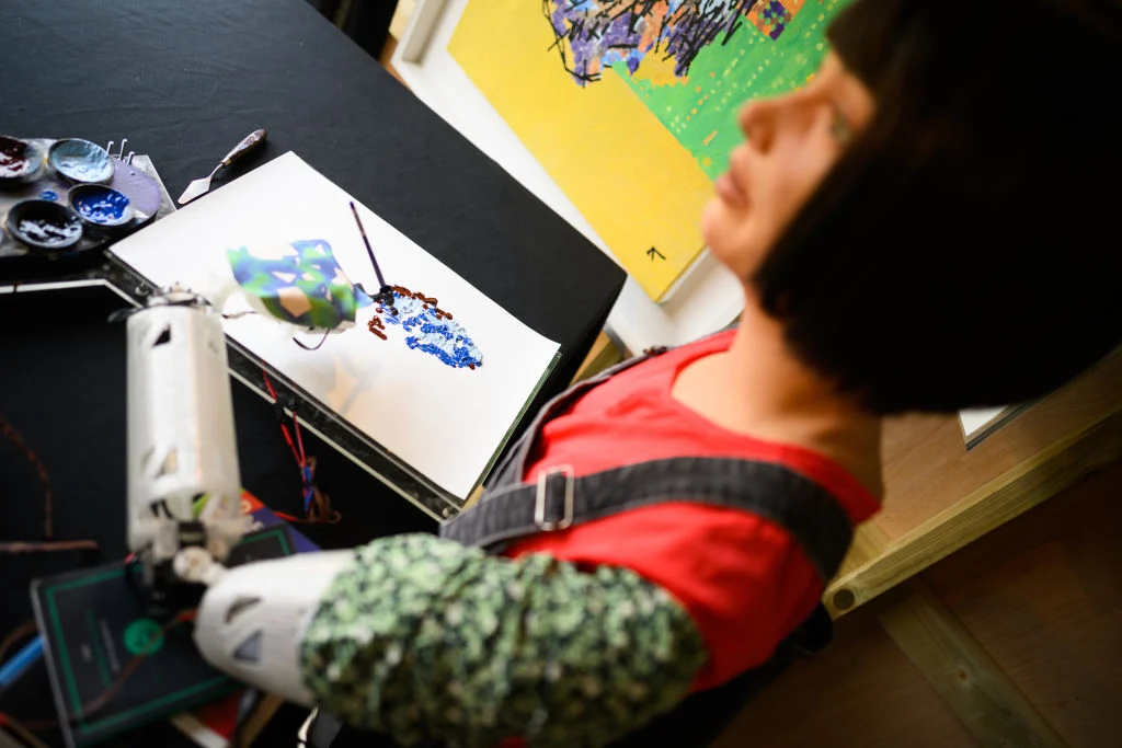 Robot o ludzkich rysach twarzy, ubrany w kolorową bluzkę i ogrodniczki, maluje na papierze abstrakcyjny obraz za pomocą sztucznej ręki, obok zestawu artystycznych farb.
