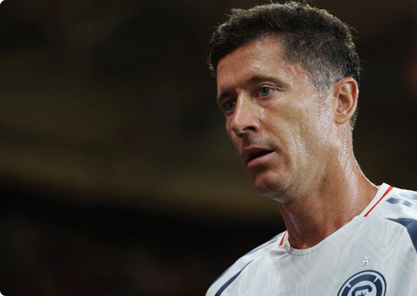 Robert Lewandowski Zbliżenie na spoconego mężczyznę w koszulce sportowej z krótkimi rękawami, skoncentrowanego podczas wysiłku fizycznego, z widocznym logotypem na piersi.