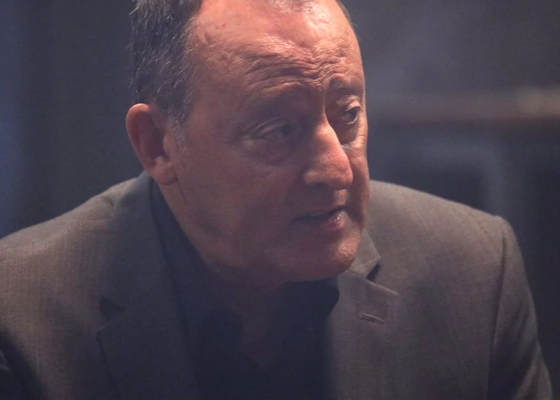 Jean Reno w filmie "Zabójcza portierka" Mężczyzna w średnim wieku w eleganckiej, ciemnej marynarce, z lekko poszarzałą cerą i poważnym wyrazem twarzy