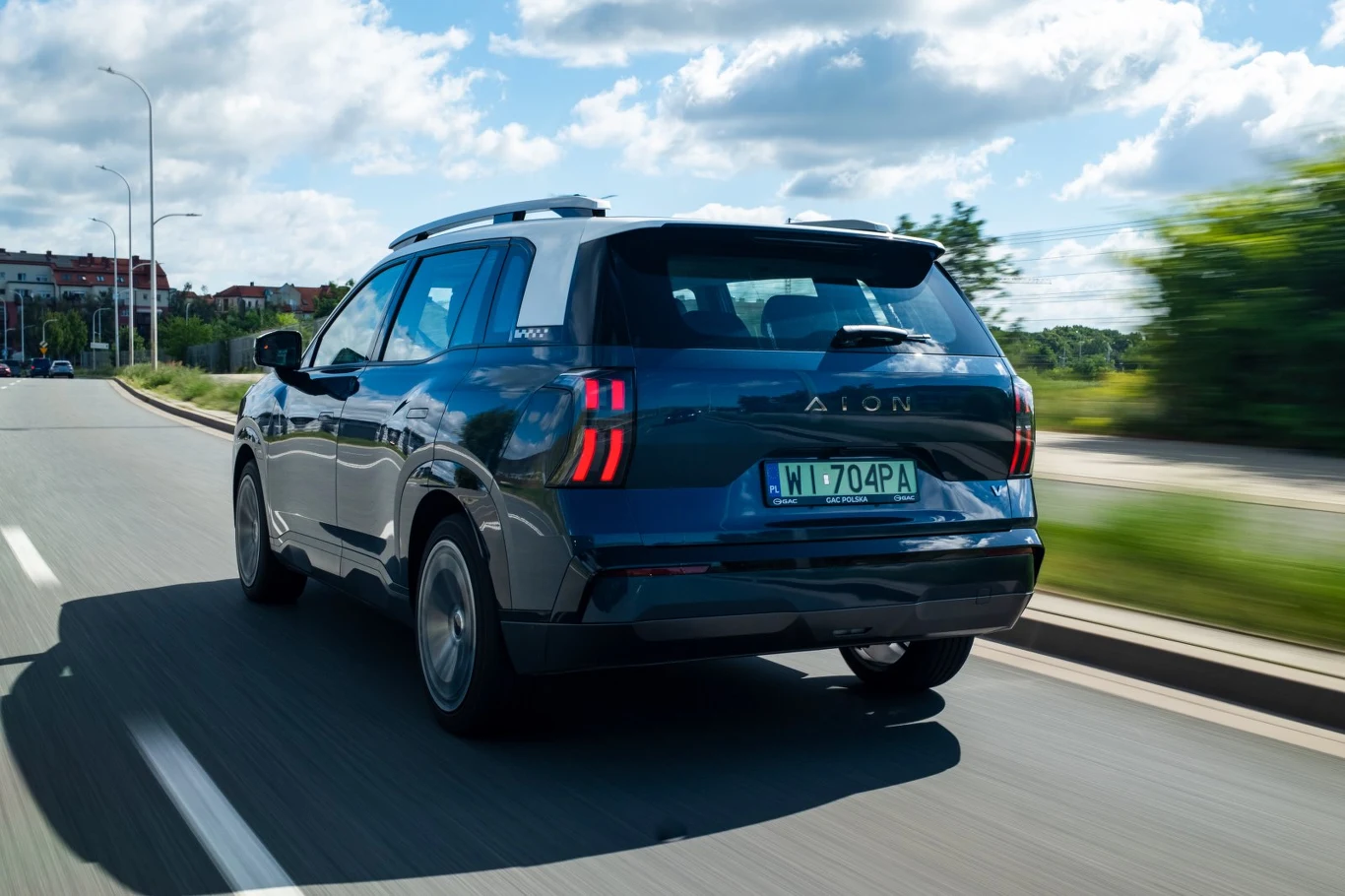 Niebieski SUV marki Aion porusza się prawym pasem miejskiej drogi w słoneczny dzień, z widocznym zielonymi tablicami rejestracyjnymi i charakterystycznymi pionowymi światłami tylnymi.