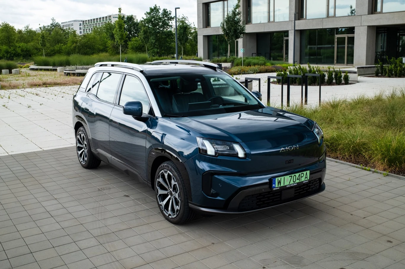 Nowoczesny, ciemnoniebieski SUV Aion stoi na placu z betonową nawierzchnią, otoczony zielenią i zabudowaniami o przeszklonych fasadach.