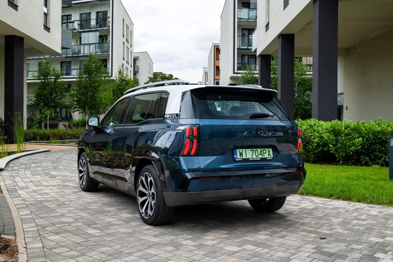 Granatowy, nowoczesny SUV zaparkowany na brukowanej alejce pomiędzy nowoczesnymi budynkami mieszkalnymi, otoczony zielenią i niskimi krzewami.