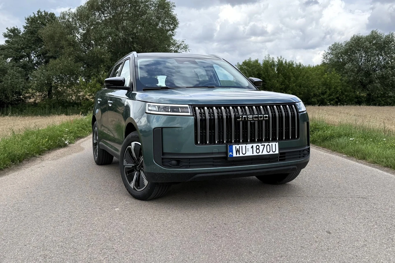Dzięki napędowi elektrycznemu hybrydowe Jaecoo 5 bardzo chętnie reaguje na gaz. Nowoczesny, zielony SUV marki Jaecoo zaparkowany na asfaltowej drodze, otoczony zielenią i polami uprawnymi, pod pochmurnym niebem.