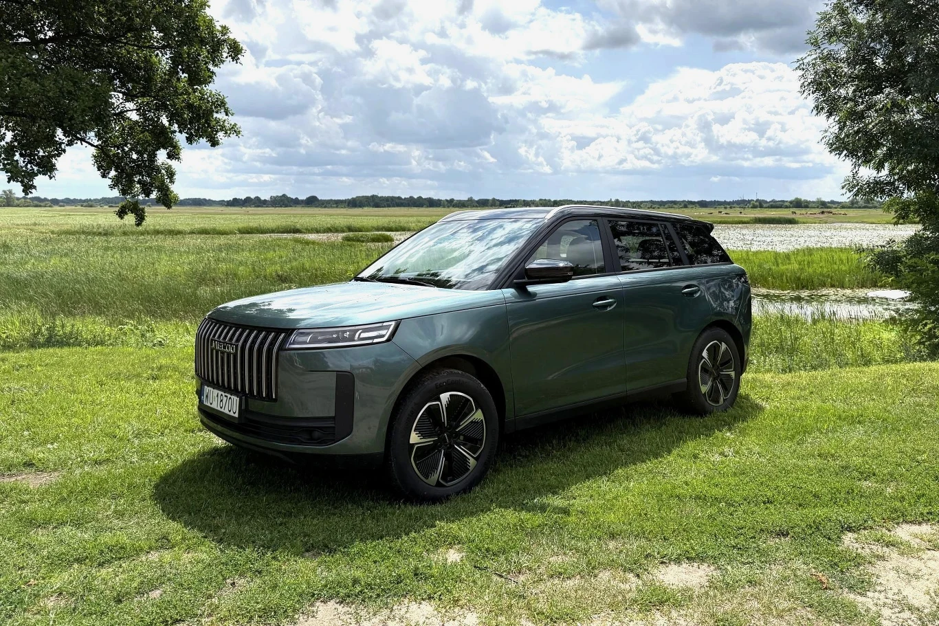 Jaecoo 5 Hybrid ma 224 KM i pali 4,3 l/100 km. Przez cenę 119 500 zł konkurenci nie mogą spać spokojnie. Zielony Jaecoo 5 Hybrid, SUV stojący na trawie przy jeziorze, widok pod zachmurzonym niebem.