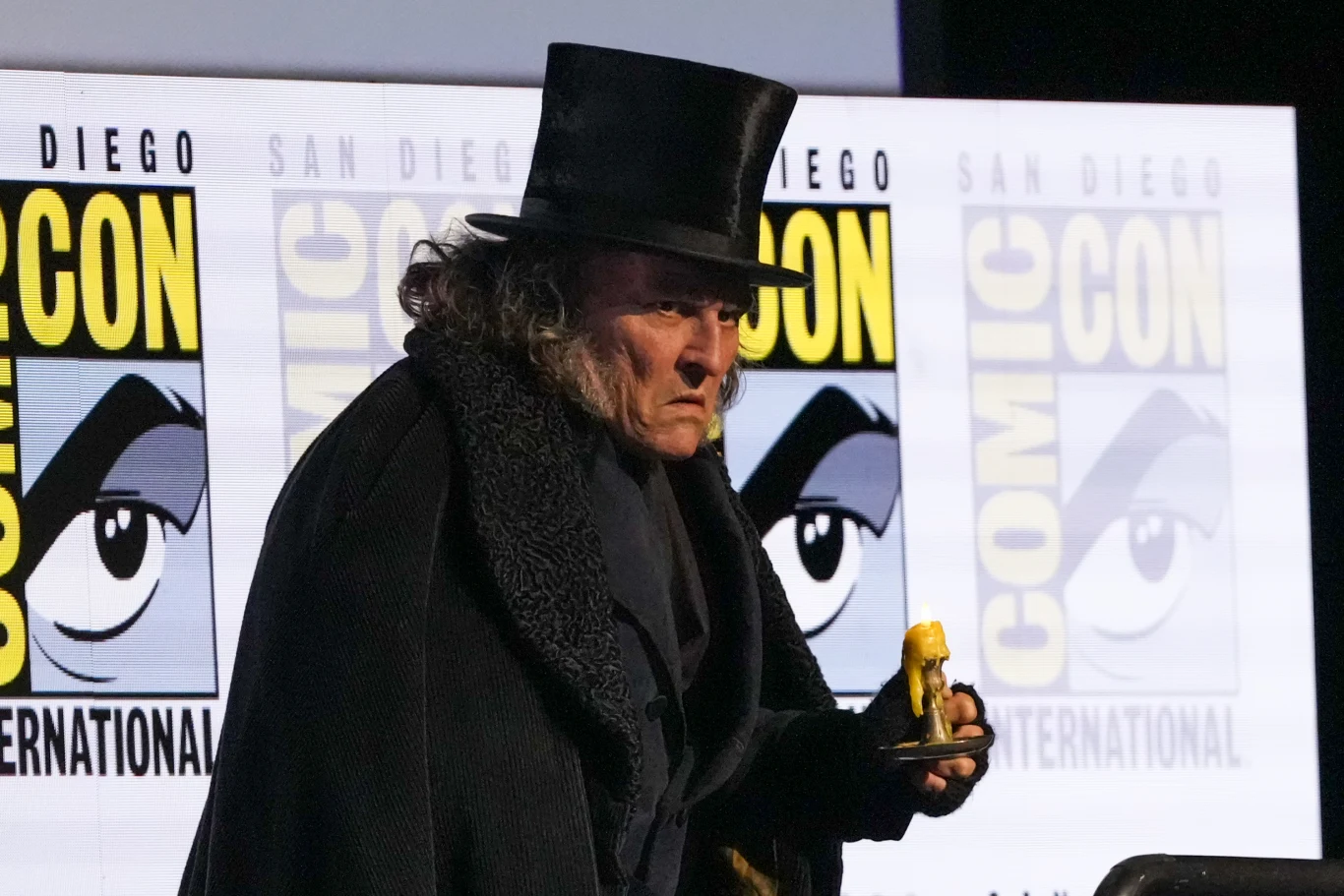 Mężczyzna w czarnym płaszczu i cylindrze, z surowym wyrazem twarzy, trzymający żółtą figurkę podczas wydarzenia na tle banera Comic Con International.