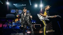 Dwóch muzyków podczas występu na scenie, wokalista w czarnej skórzanej kurtce i czapce śpiewa, a gitarzysta w żółtym stroju energicznie gra na gitarze, w tle perkusja i ekrany świetlne podkreślają koncertową atmosferę.