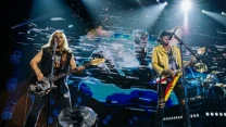 Dwóch muzyków grających na gitarach, dynamiczna scenografia z efektami świetlnymi i nowoczesną wizualizacją w tle, perkusista widoczny w dalszym planie.