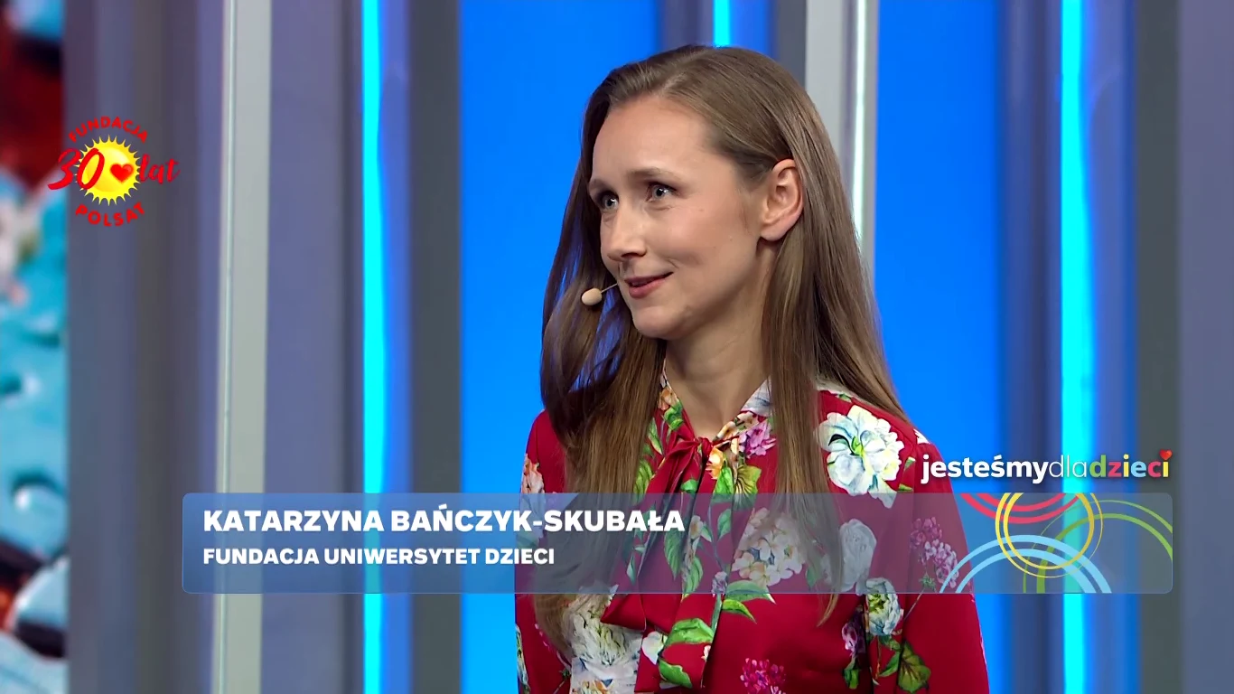 Kobieta o długich, jasnobrązowych włosach w czerwonej, kwiecistej bluzce pozuje na tle niebieskiej scenografii telewizyjnej z widocznymi grafikami i napisem „jesteśmy dzieci”.