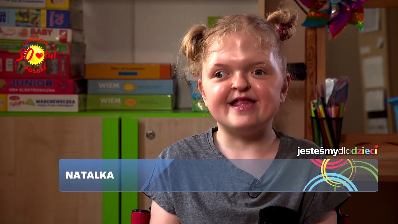 Uśmiechnięta dziewczynka z jasnymi włosami spiętymi w dwa kucyki siedzi na tle regału z kolorowymi grami planszowymi, po lewej stronie na ekranie widoczna jest jej imię – Natalka, a w prawym górnym rogu znajdują się kolorowe grafiki i logo kampanii 'je...