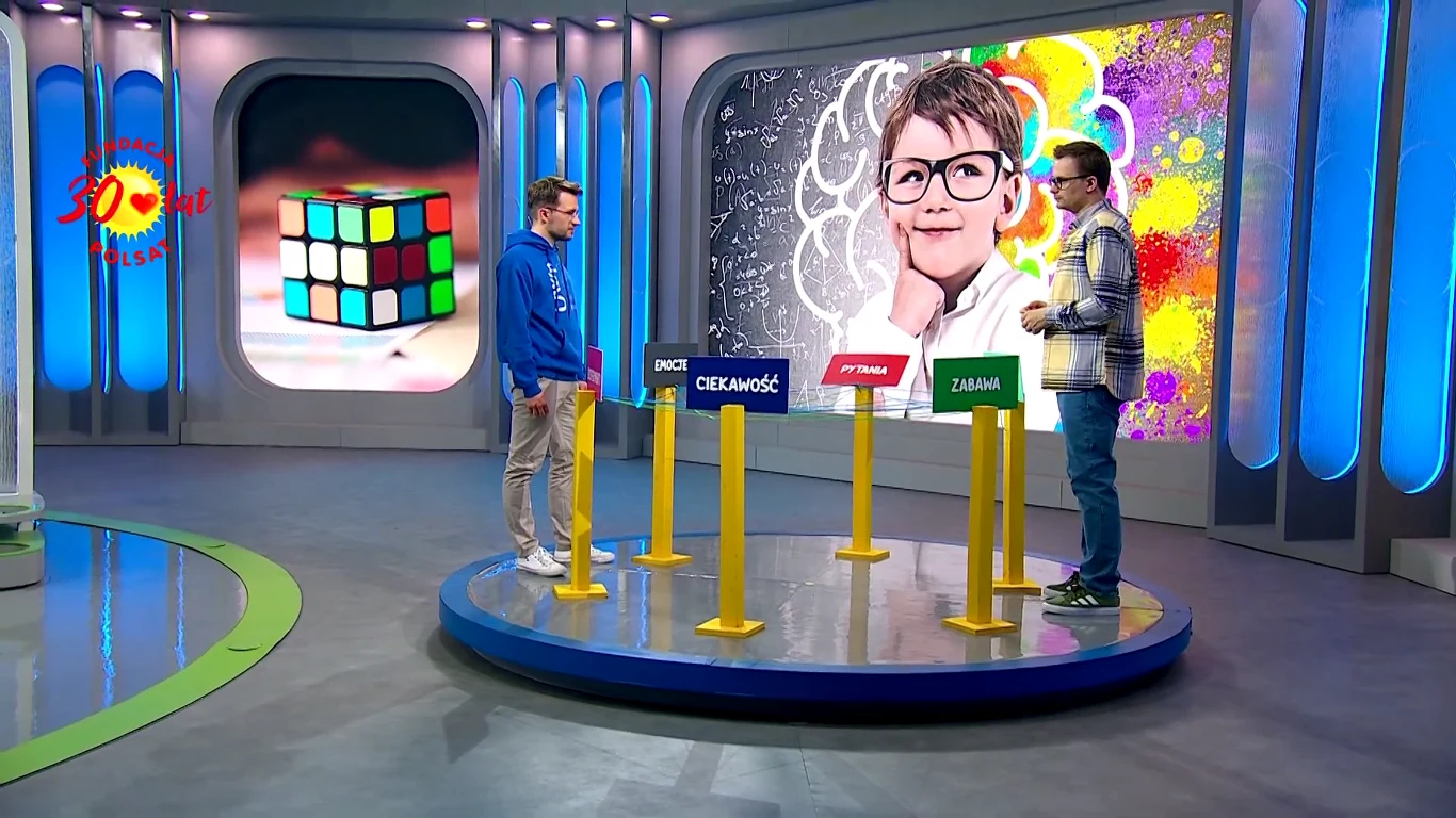 Dwóch mężczyzn rozmawia i wskazuje na tabliczki z napisami „Pasja”, „Ciekawość”, „Zabawa”, stojące na okrągłej scenie, za nimi kolorowa ilustracja dziecka z okularami oraz podświetlona kostka Rubika w tle.
