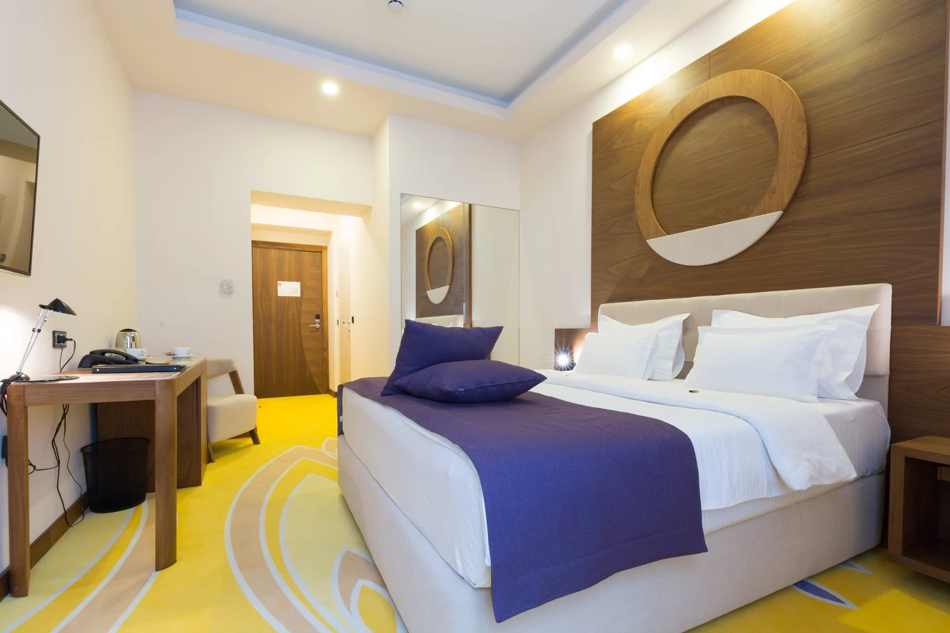 Nowoczesny pokój hotelowy z dużym łóżkiem, fioletową narzutą i poduszkami, ozdobną ścianą z drewnianym okręgiem, biurkiem po lewej stronie oraz lustrem naprzeciwko łóżka.
