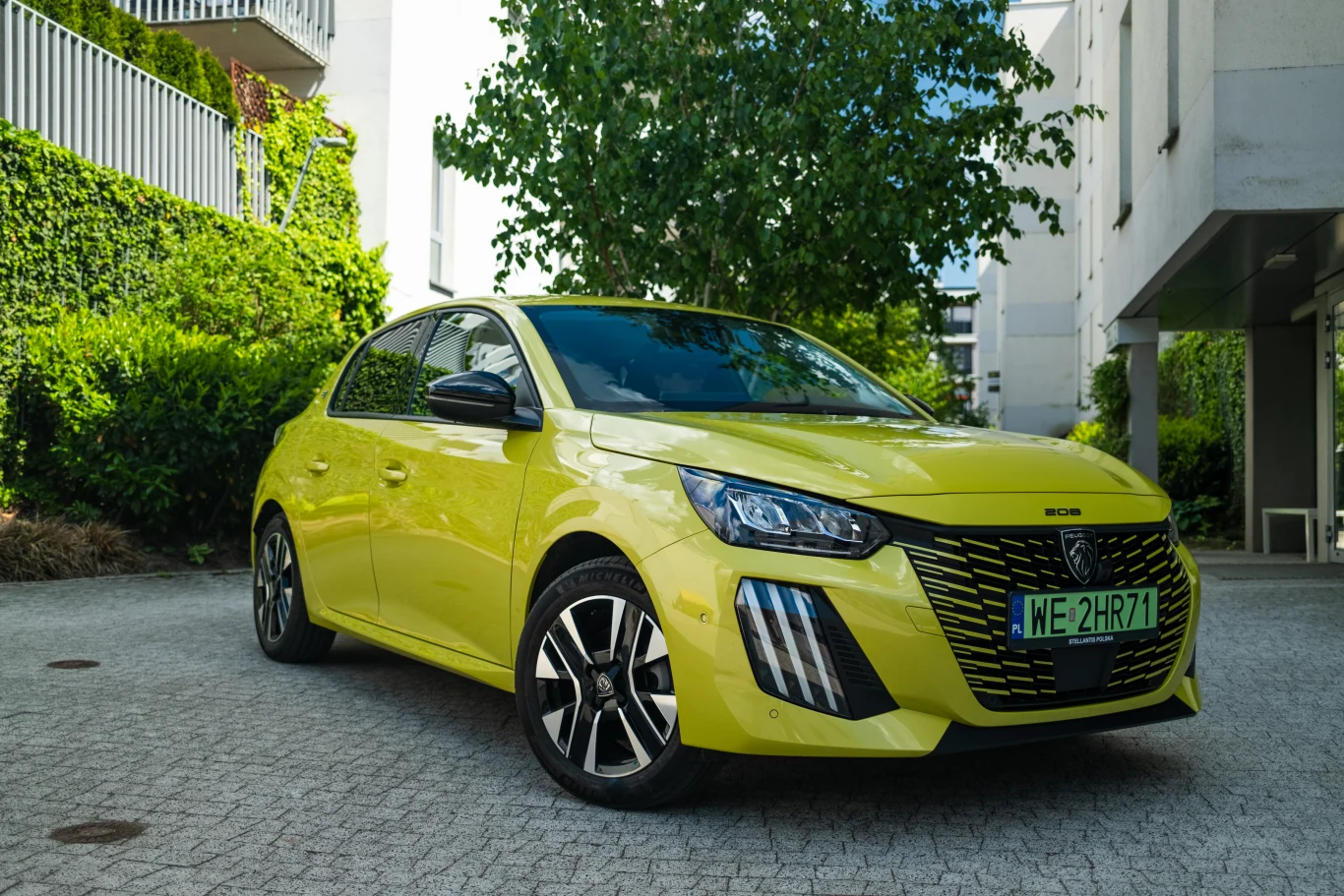 Jasnożółty, nowoczesny hatchback Peugeot 208 stoi zaparkowany na dziedzińcu otoczonym zielenią i zabudową mieszkalną.