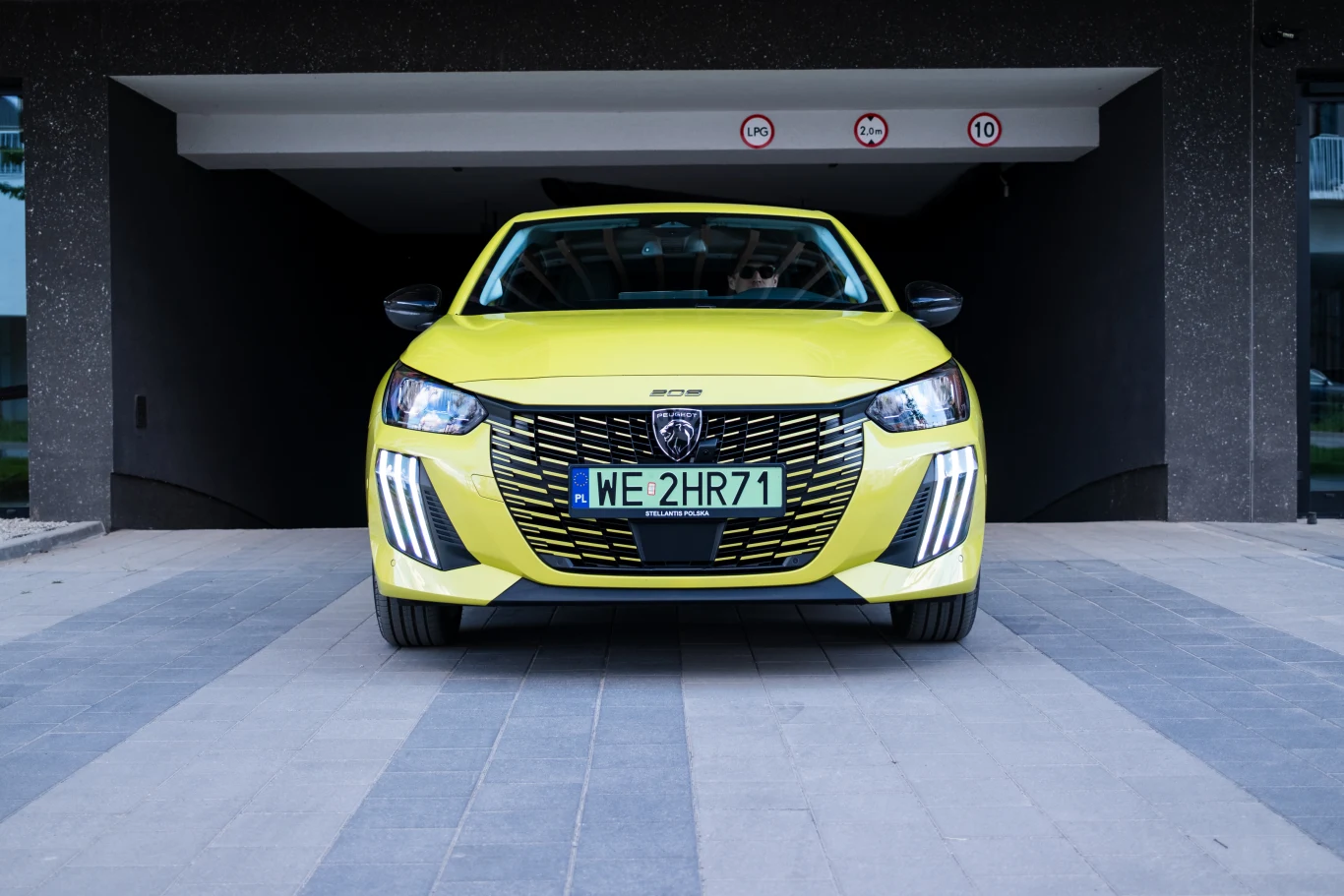 Żółty samochód osobowy Peugeot stoi przodem do obiektywu przed wjazdem do garażu, z widoczną tablicą rejestracyjną WE 2HR71 i charakterystycznym nowoczesnym grillem.
