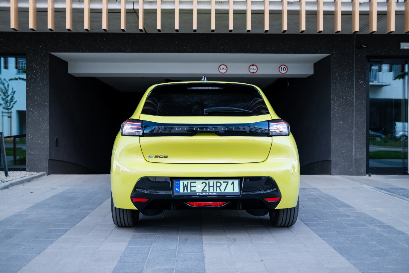 Jaskrawożółty samochód Peugeot e-208 ustawiony tyłem przed wjazdem do ciemnego garażu, z widoczną tablicą rejestracyjną WE 2HR71 i czarnym tylnym zderzakiem.